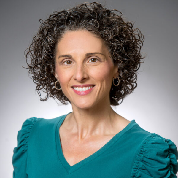 Mindy L. Calandro, wearing a teal top, smiles warmly at the camera, her curly hair framing her face against a neutral background.