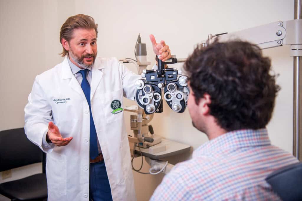 An optometry doctor in a white coat explains a phoropter to a patient during an eye exam in a clinic room.