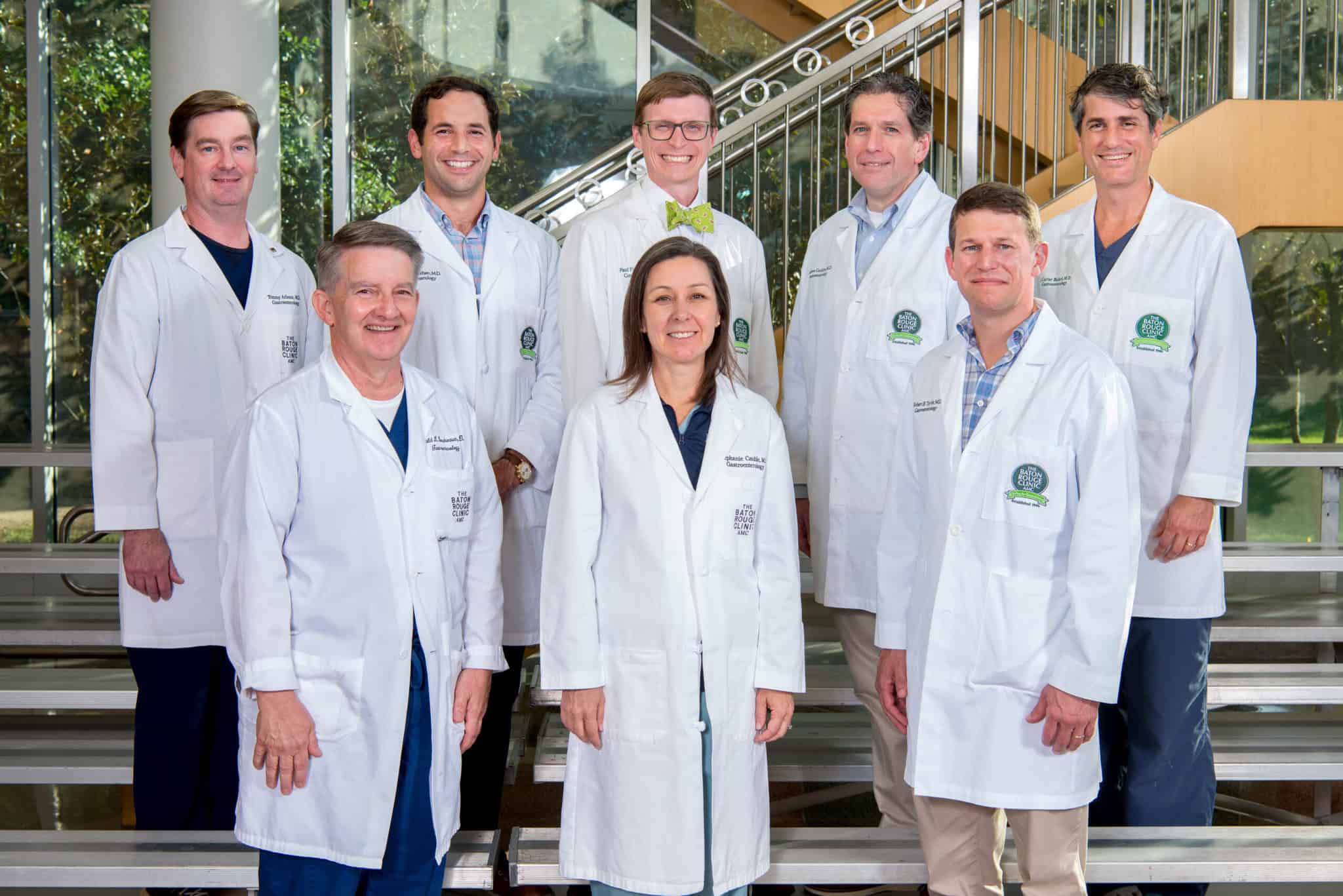A team of eight gastroenterology experts, seven men and one woman, in white coats gather on stairs in a brightly lit indoor setting.