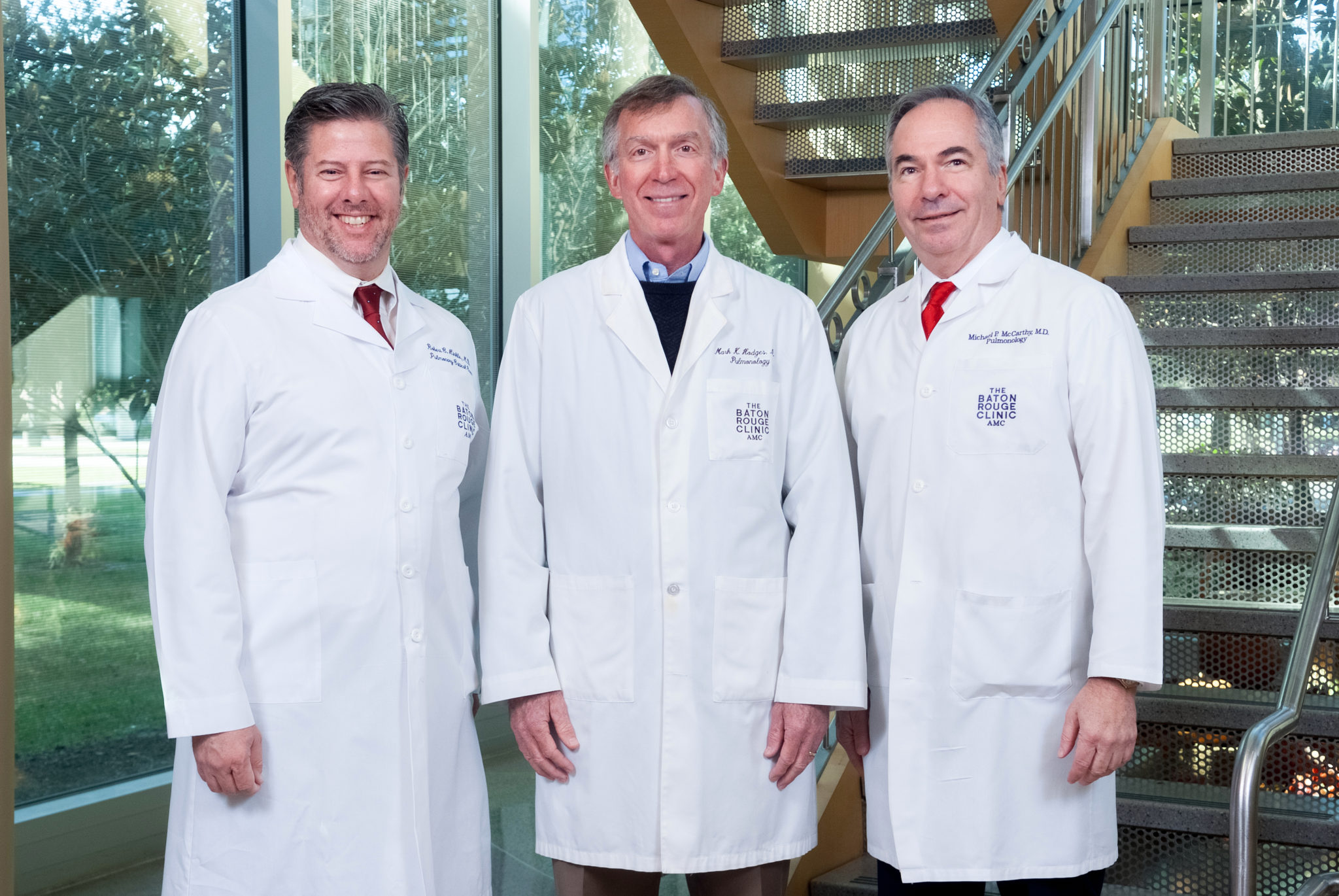 Three men in white lab coats, physicians from The Baton Rouge Clinic, stand side by side near a staircase in a building with large windows.