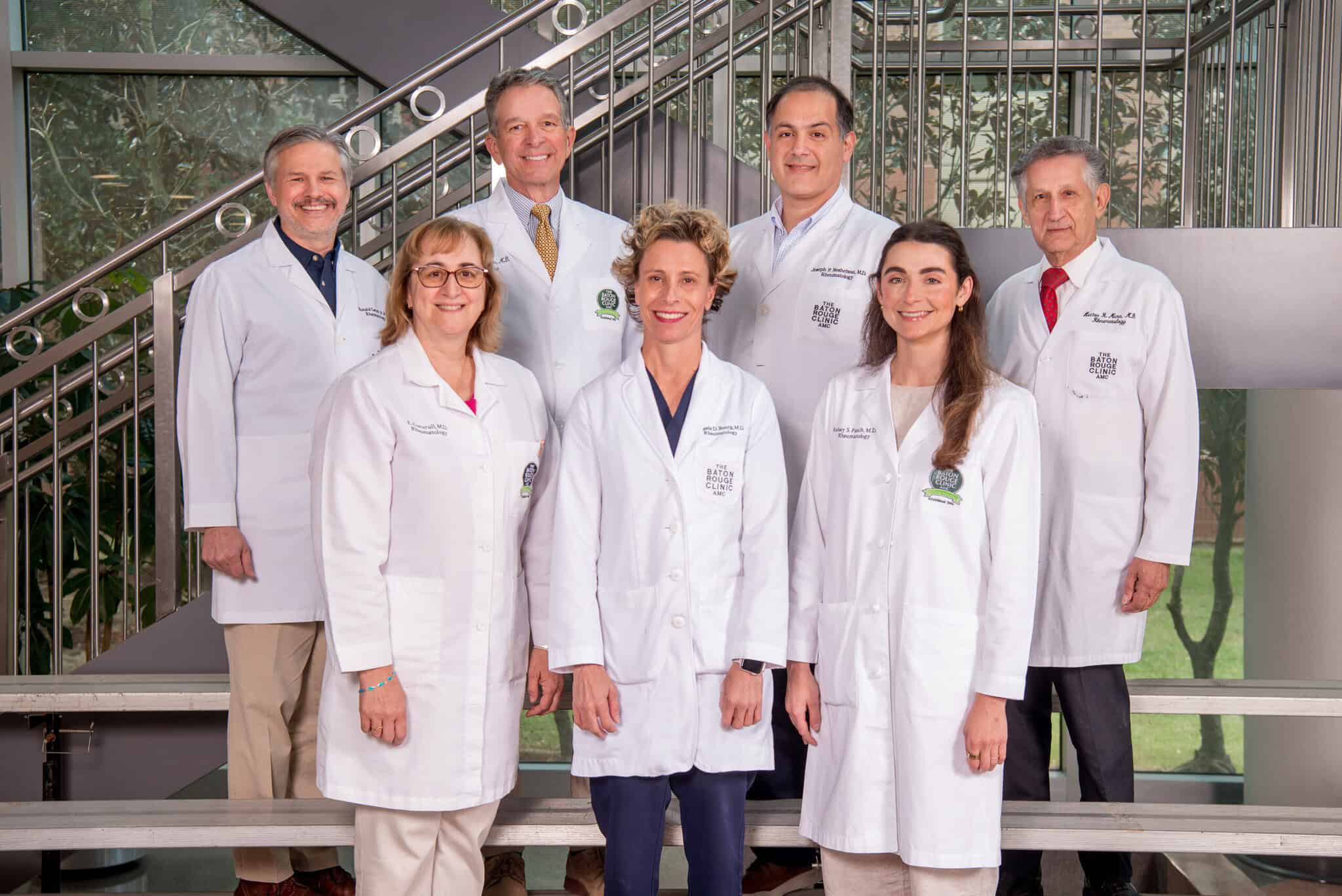 Seven adults in white lab coats, likely part of a rheumatology team, pose indoors for a group photo by a staircase and windows.