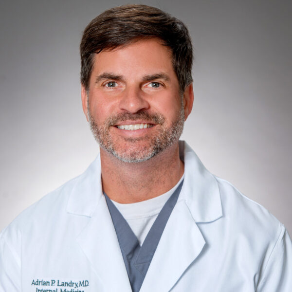 Adrian P. Landry, in a white medical coat with "Internal Medicine" embroidered, smiles at the camera against a neutral grey background.