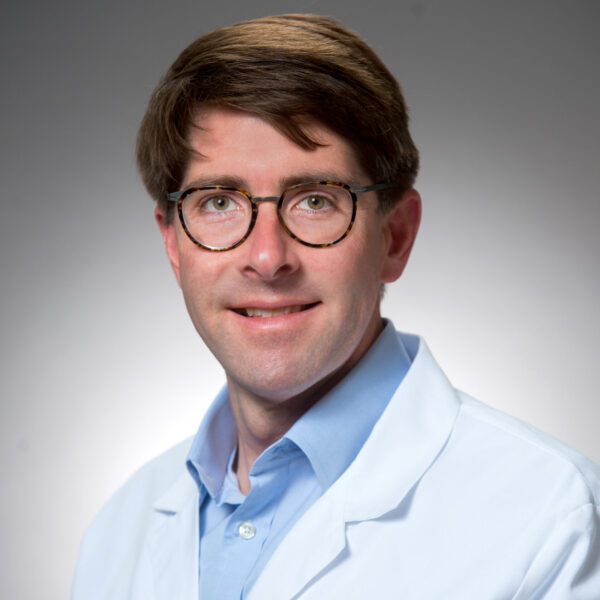 Dr. Stephen Sanches, in glasses and a white coat adorned with medical titles, poses confidently against a gray background.