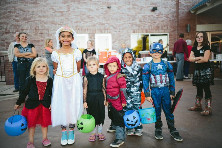 Kids in Halloween costumes with trick-or-treat buckets gather outside a brick building, as Louisiana doctors chat at tables in the background.