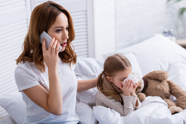mother calling doctor by smartphone, sick daughter blowing nose in bed