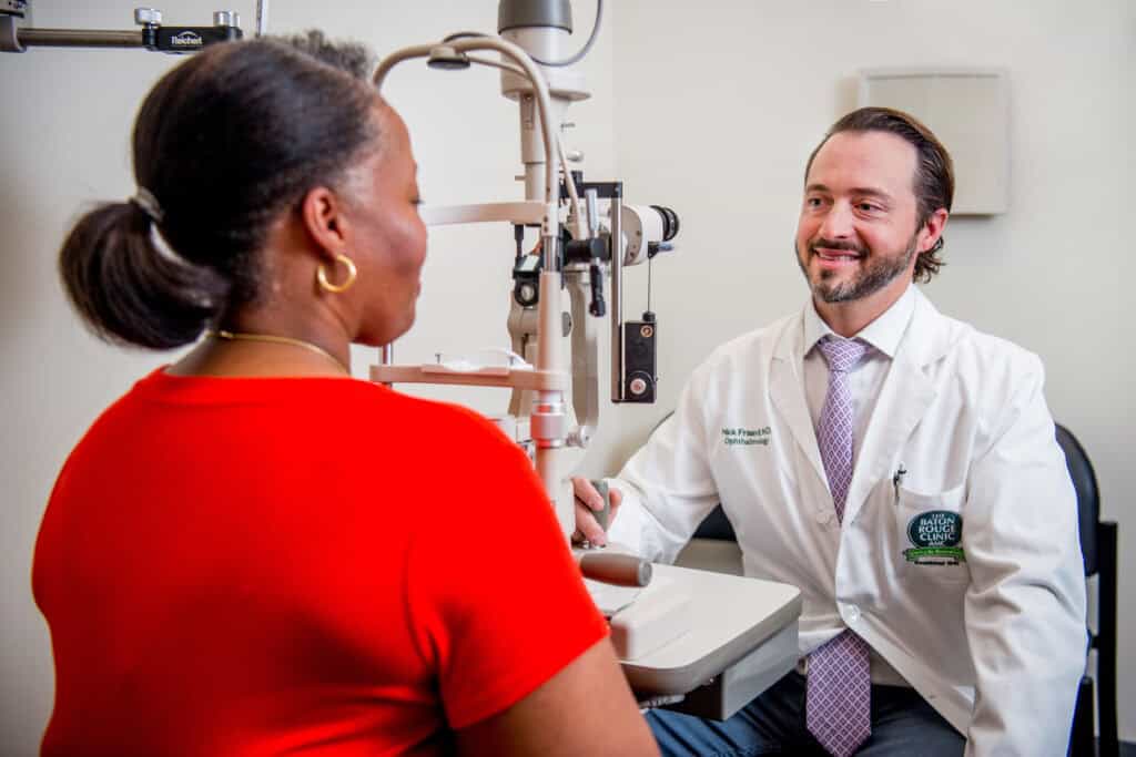 A doctor in a white coat speaks to a patient in a red shirt during an eye exam at a medical office.