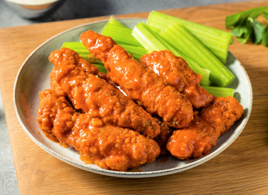 A plate of buffalo chicken tenders with celery sticks, served on a wooden board with a cup of ranch dressing in the background.