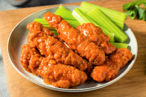 A plate of buffalo chicken tenders with celery sticks, served on a wooden board with a cup of ranch dressing in the background.