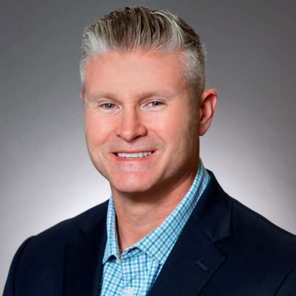A man with short gray hair in a navy suit jacket and blue checkered shirt poses in front of a plain gray background.