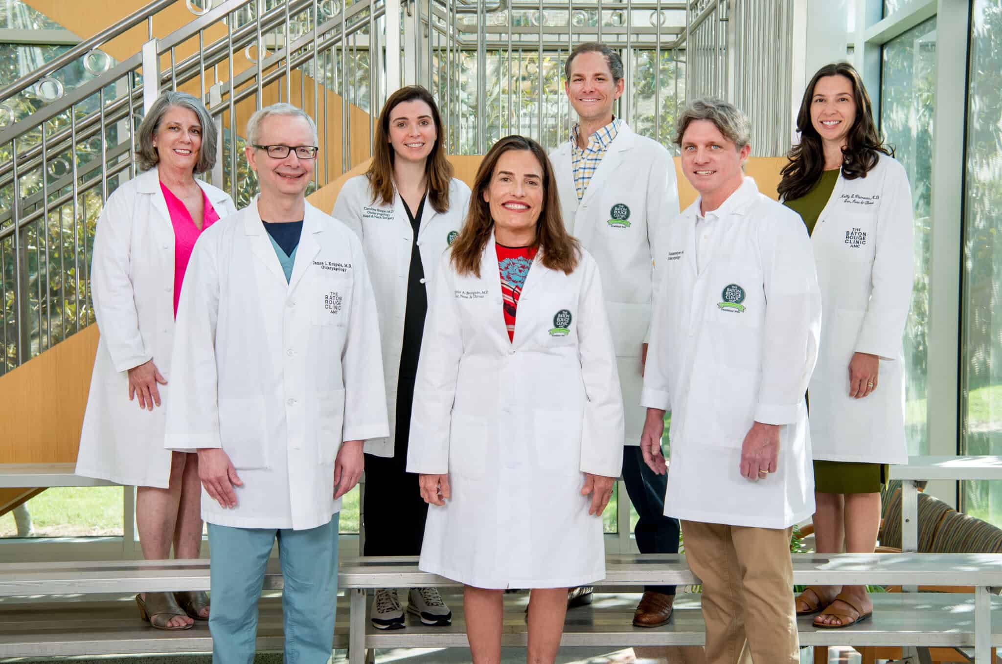 Seven people in white lab coats stand and smile together in a bright, modern indoor setting with metal railing and large windows.