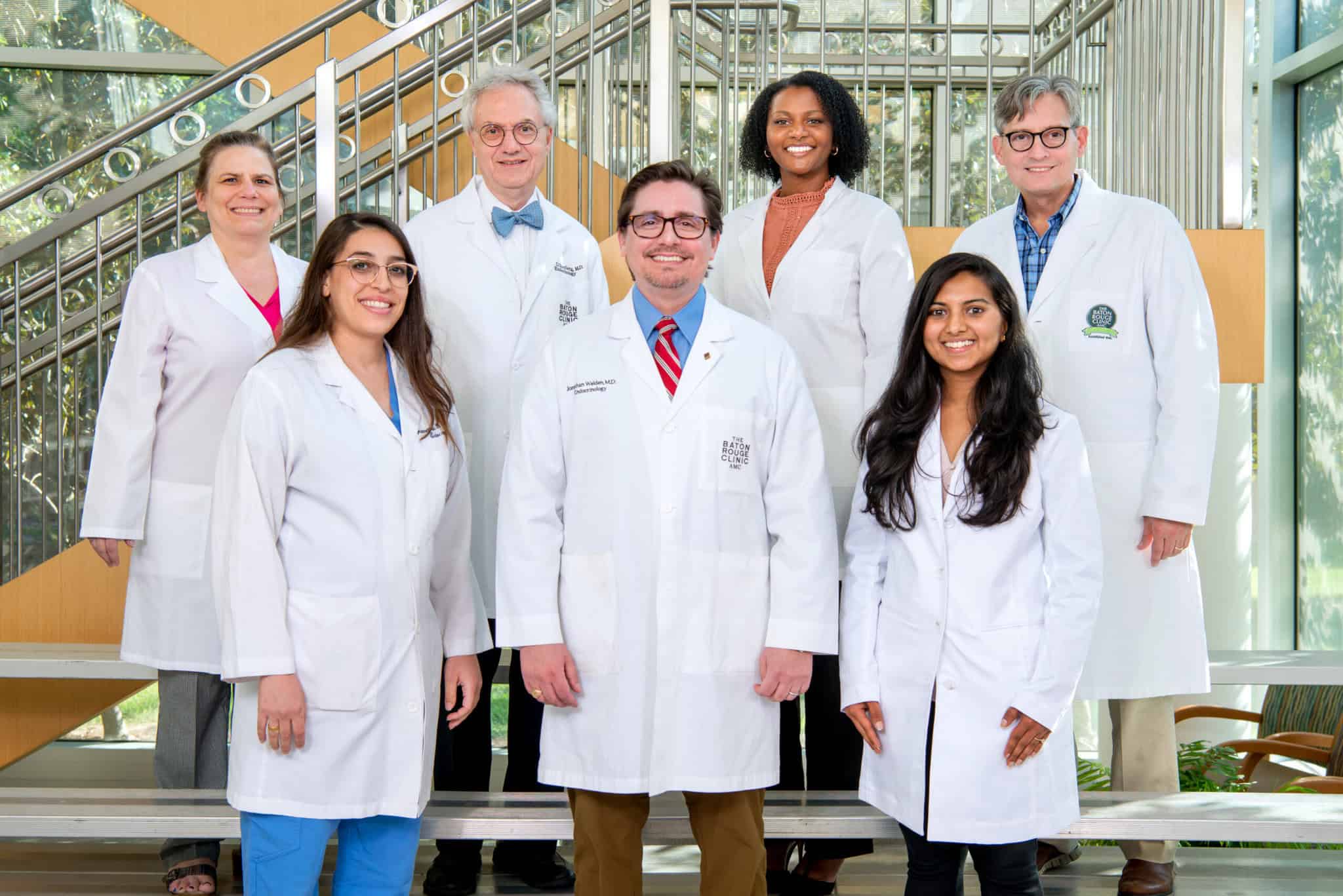 Seven people in white lab coats stand and smile on an indoor staircase with glass walls and natural light in the background.