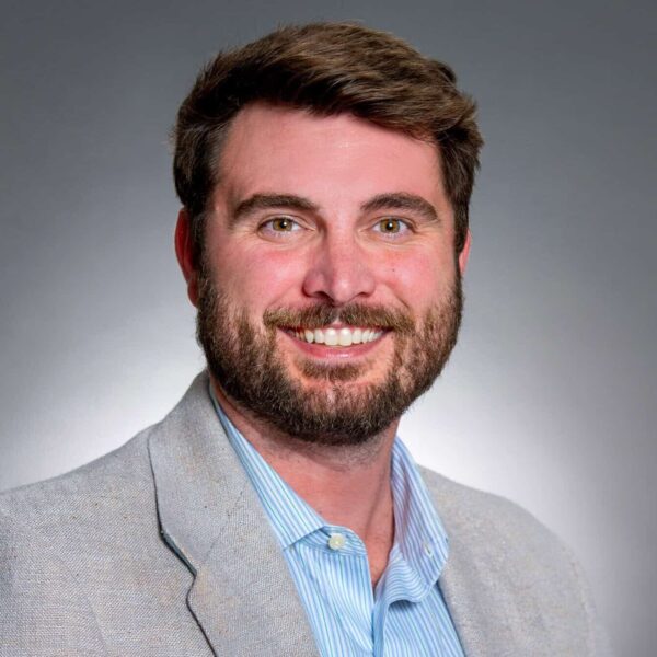 A man with brown hair and a beard wearing a light gray blazer and blue striped shirt poses in front of a gray background.