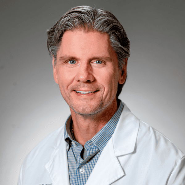 A middle-aged man with gray hair in a white lab coat and checked shirt, smiling, in front of a plain gray background.