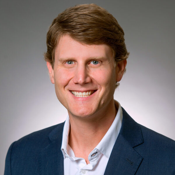 Smiling man in a blue suit jacket and white shirt, facing the camera against a plain, light gray background.