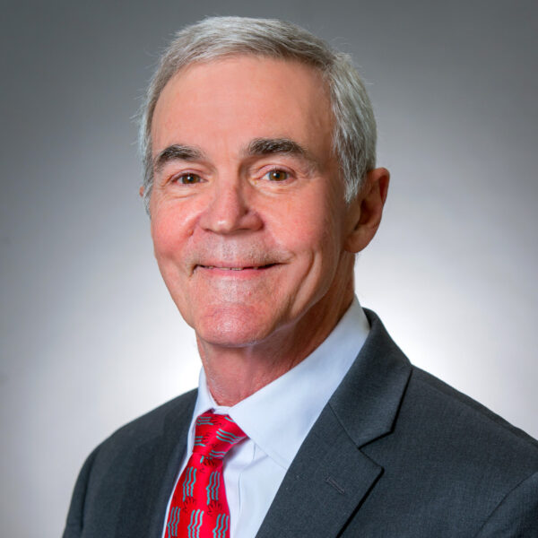 Older man in a gray suit, light blue shirt, and red patterned tie, smiling at the camera against a plain gray background.