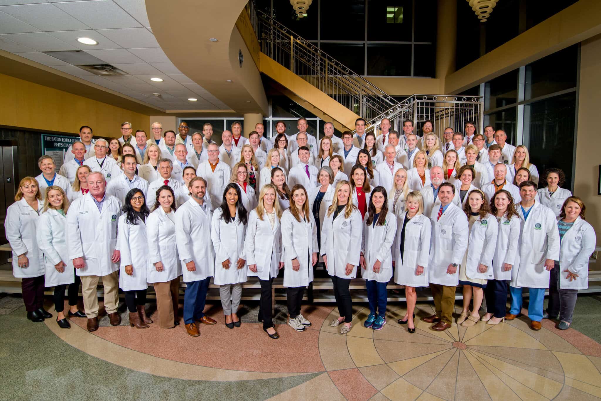 A large group of Louisiana doctors in white lab coats stands together in a spacious lobby with a staircase in the background.