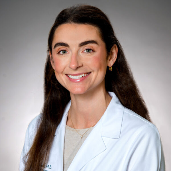 A woman with long brown hair wearing a white medical coat, smiling in front of a plain gray background.