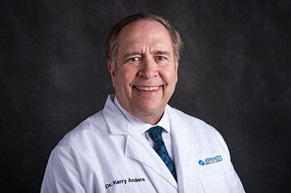 Smiling man in a white lab coat with a blue patterned tie, representing Baton Rouge Physicians, against a dark background.