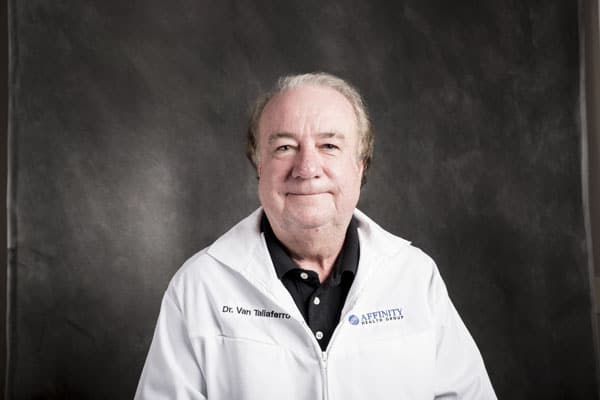 An older man with light hair, donning a white jacket and black shirt, stands poised against a dark gray background at The Baton Rouge Clinic.