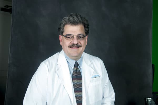 Man with glasses and a mustache, in a white lab coat and patterned tie, embodies expertise at The Baton Rouge Clinic against a dark background.