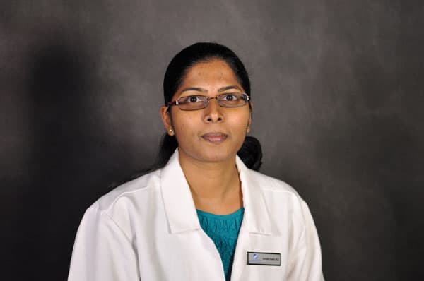 Person with glasses wearing a white lab coat and blue shirt, resembling a Baton Rouge physician, against a dark grey background.