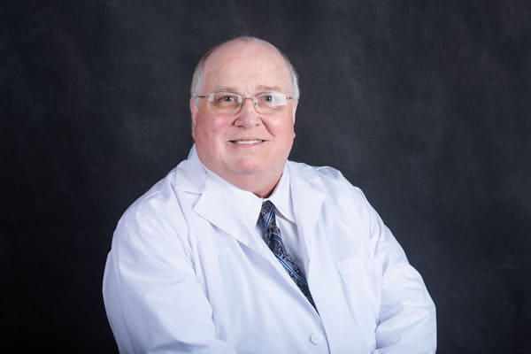 A Baton Rouge Clinic doctor in a white lab coat and patterned tie smiles against a dark background.