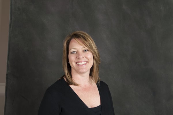 A person with shoulder-length hair smiling, wearing a black top, stands in front of a gray studio backdrop at The Baton Rouge Clinic.