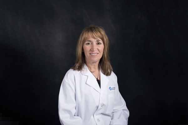 A person with shoulder-length hair, wearing a white coat, stands against a dark background, embodying the expertise of Baton Rouge Physicians.