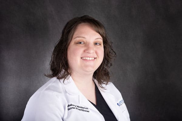 A person with shoulder-length brown hair, wearing a white lab coat and a black top, is smiling at The Baton Rouge Clinic against a dark gray background.