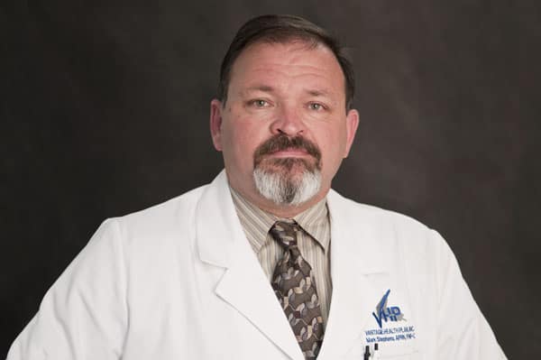 Man with a beard in a white lab coat and patterned tie, ready to provide expert care at The Baton Rouge Clinic against a dark background.