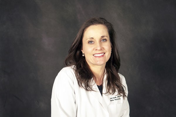 A woman with long brown hair smiles in a white lab coat, representing the skilled Baton Rouge Physicians against a dark grey background.