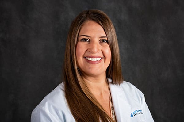 Person with long brown hair, wearing a light blue lab coat, smiles against a dark gray background, representing Baton Rouge Physicians.