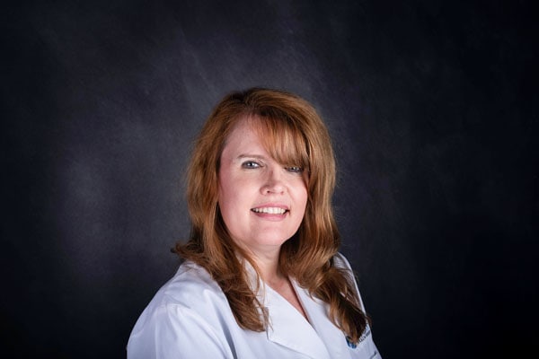 A woman with long brown hair, from The Baton Rouge Clinic, smiles in a white lab coat against a dark gray background.