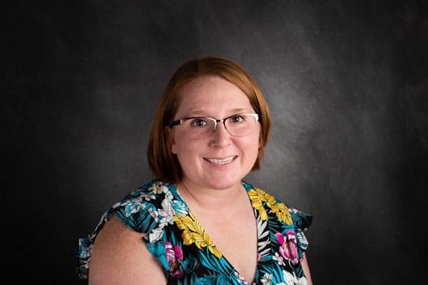 A woman wearing glasses and a floral top smiles against a dark background, embodying the care of Baton Rouge physicians.