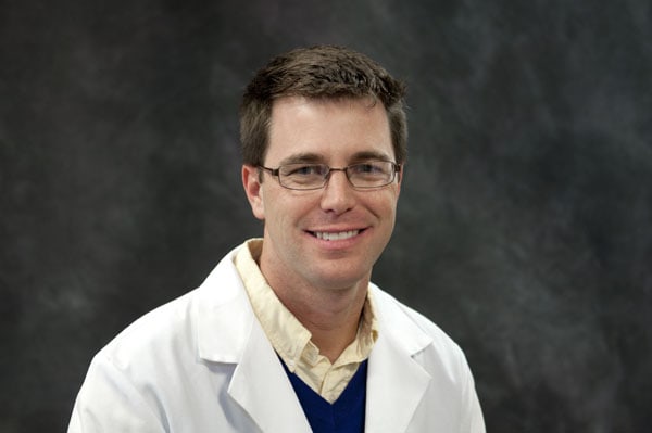 A Baton Rouge physician in glasses and a lab coat smiles against a dark studio background.