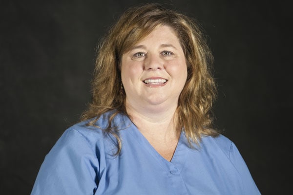 A woman with long brown hair in a blue scrub shirt, smiling warmly as if embodying care at The Baton Rouge Clinic.