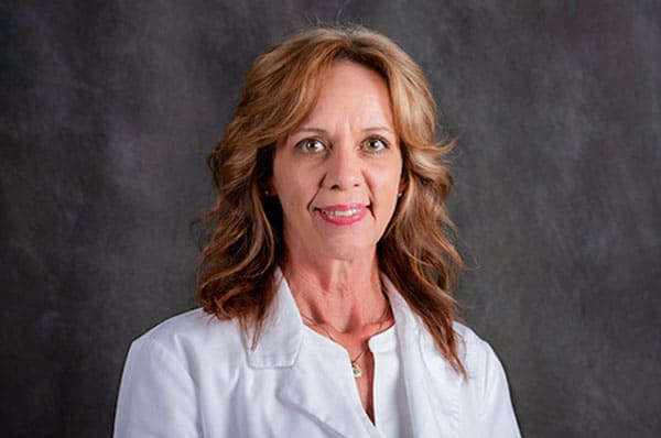 Woman with wavy light brown hair and a white coat stands against a dark gray background, facing forward and smiling slightly; Baton Rouge physician.