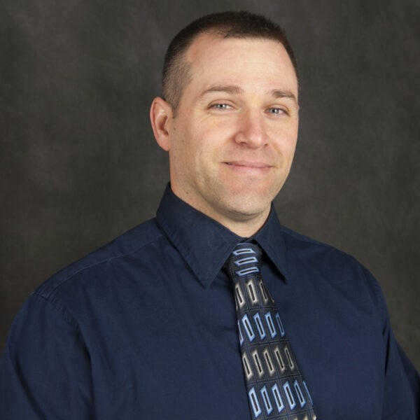 A man wearing a dark blue shirt and patterned tie, reminiscent of Baton Rouge physicians, stands against a dark background with a slight smile.