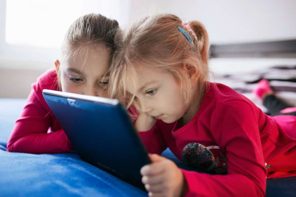Two young children lie on a blue blanket in Baton Rouge, intently looking at a tablet. Both are wearing pink shirts and have their hair tied back.