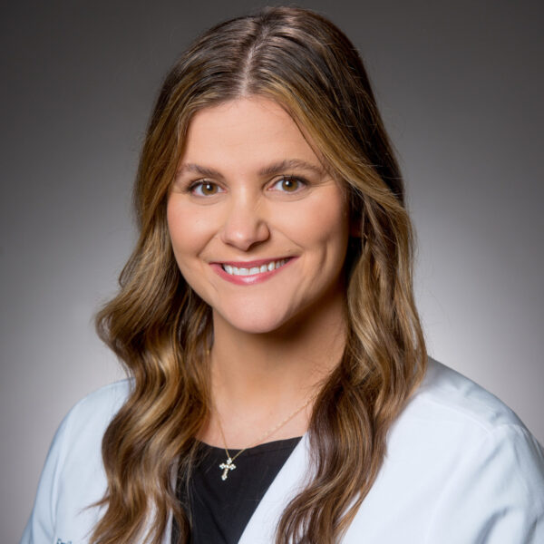 A woman with wavy brown hair wearing a white lab coat and black top, smiling in front of a gray background.