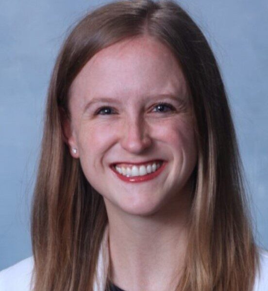 A woman with long hair smiles, wearing a white lab coat against a light blue background.