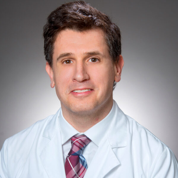 A man in a white coat with a personalized name, wearing a checkered tie, is looking at the camera against a plain background.