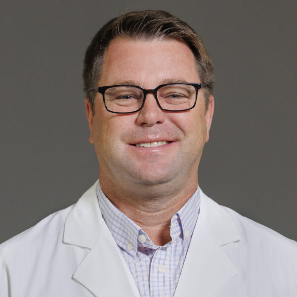 A man wearing glasses and a white medical coat with "Stuart L. Melton, M.D. Family Medicine" embroidered on it, against a gray background.