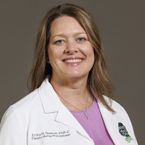 A woman in a white medical coat over a lavender shirt smiles at the camera against a plain background.