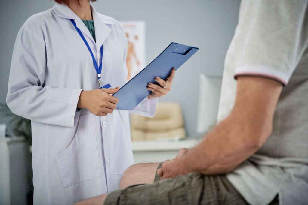 A doctor holding a clipboard speaks with a seated patient in a medical office.