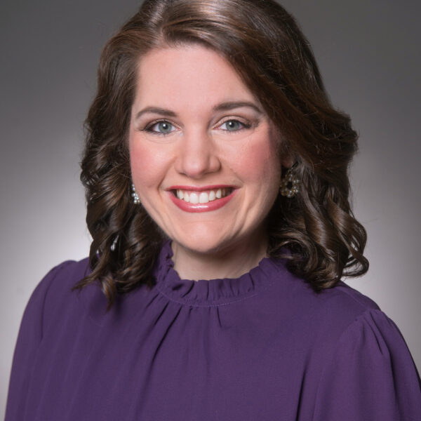 Woman with shoulder-length brown hair wearing a purple blouse, smiling in front of a plain gray background.