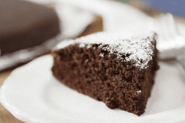 A slice of chocolate cake dusted with powdered sugar on a white plate, with a fork beside it. The rest of the cake is in the background.