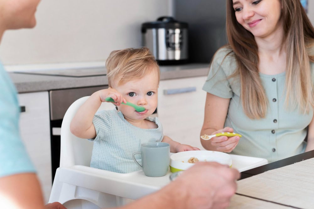 A baby sits in a high chair eating with a spoon, while two adults sit nearby at a kitchen table, one smiling.