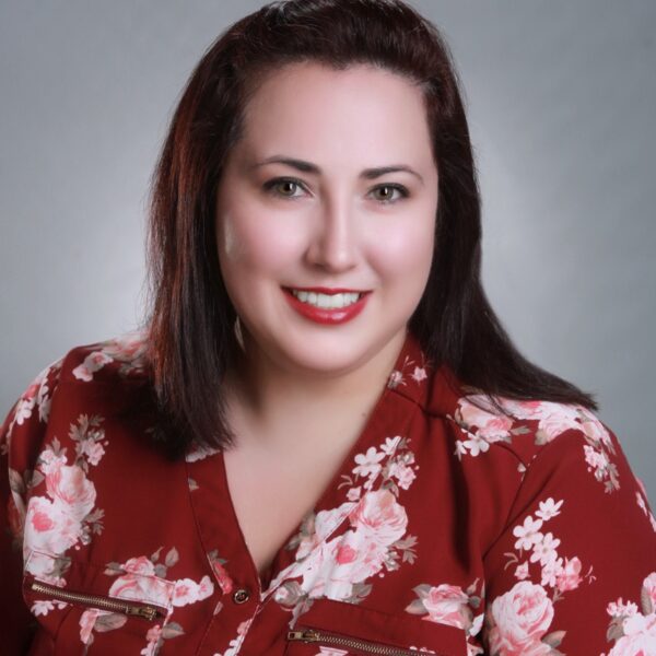 A woman with straight, dark hair wears a red floral blouse and smiles at the camera against a plain gray background.