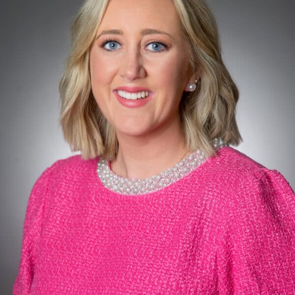 A woman with blonde hair wearing a bright pink textured top and pearl necklace, smiling in front of a gray background.